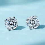 A pair of round-cut clear gemstone stud earrings in a four-prong sterling silver setting, displayed against a soft blue background