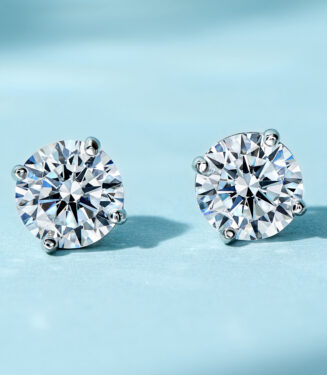 A pair of round-cut clear gemstone stud earrings in a four-prong sterling silver setting, displayed against a soft blue background