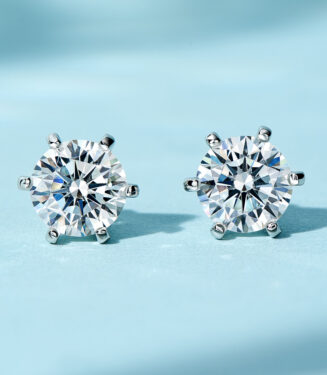 A pair of round-cut clear gemstone stud earrings in a six-prong sterling silver setting, displayed against a soft blue background.