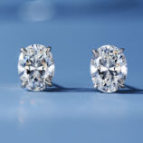 A pair of oval-cut clear gemstone stud earrings in a four-prong sterling silver setting, displayed against a soft blue background.