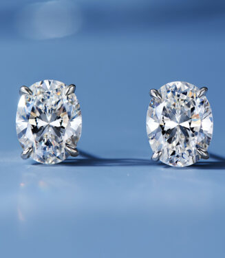 A pair of oval-cut clear gemstone stud earrings in a four-prong sterling silver setting, displayed against a soft blue background.