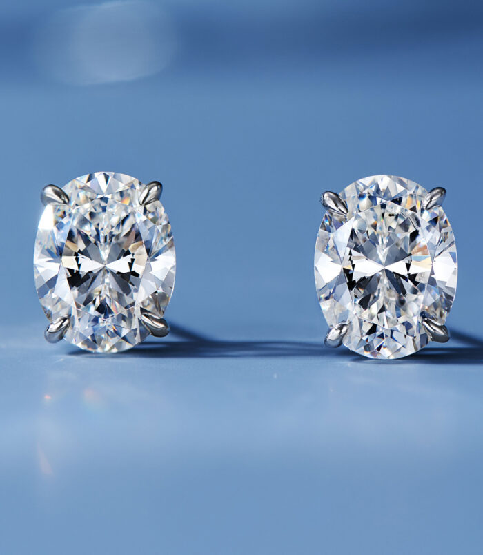 A pair of oval-cut clear gemstone stud earrings in a four-prong sterling silver setting, displayed against a soft blue background.