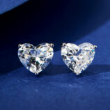 A pair of heart-shaped clear gemstone stud earrings in a four-prong sterling silver setting, displayed against a deep blue velvet background.