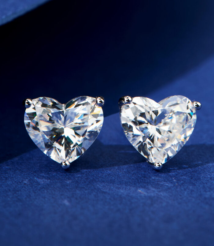 Romantic Heart-Shaped Sterling Silver Stud Earrings A pair of heart-shaped clear gemstone stud earrings in a four-prong sterling silver setting, displayed against a deep blue velvet background.