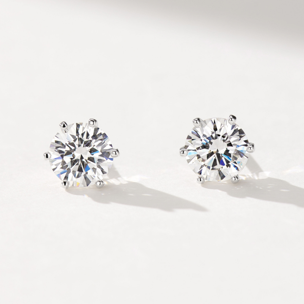 A pair of round, brilliant-cut stud earrings in polished sterling silver, featuring a central clear gemstone set in a classic six-prong setting, displayed against a soft white background.