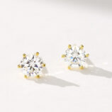 A pair of round, brilliant-cut stud earrings in polished sterling silver, featuring a central clear gemstone set in a classic six-prong setting, displayed against a soft white background.