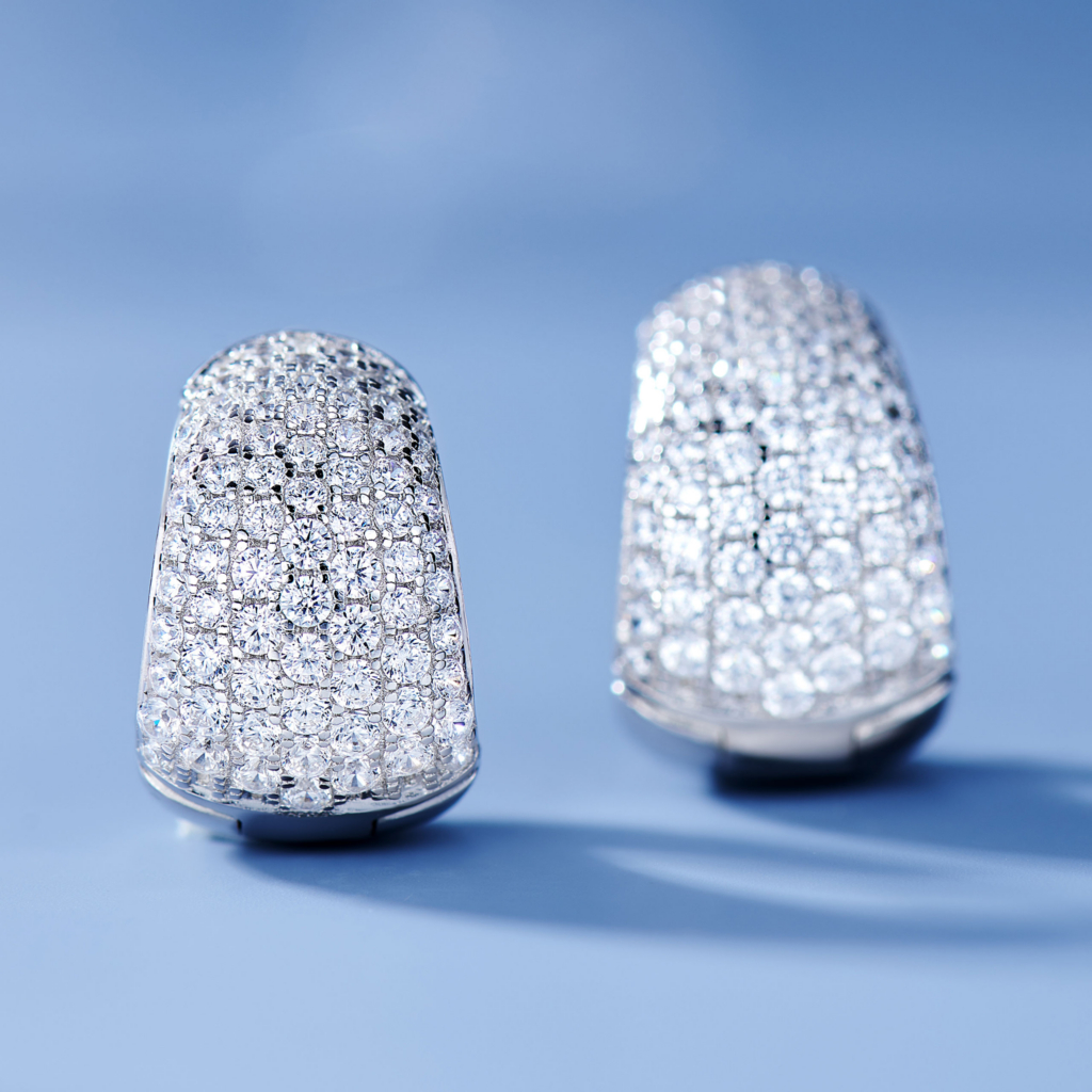 A pair of sparkling sterling silver earrings covered in small, round, clear gemstones, set against a soft blue background.