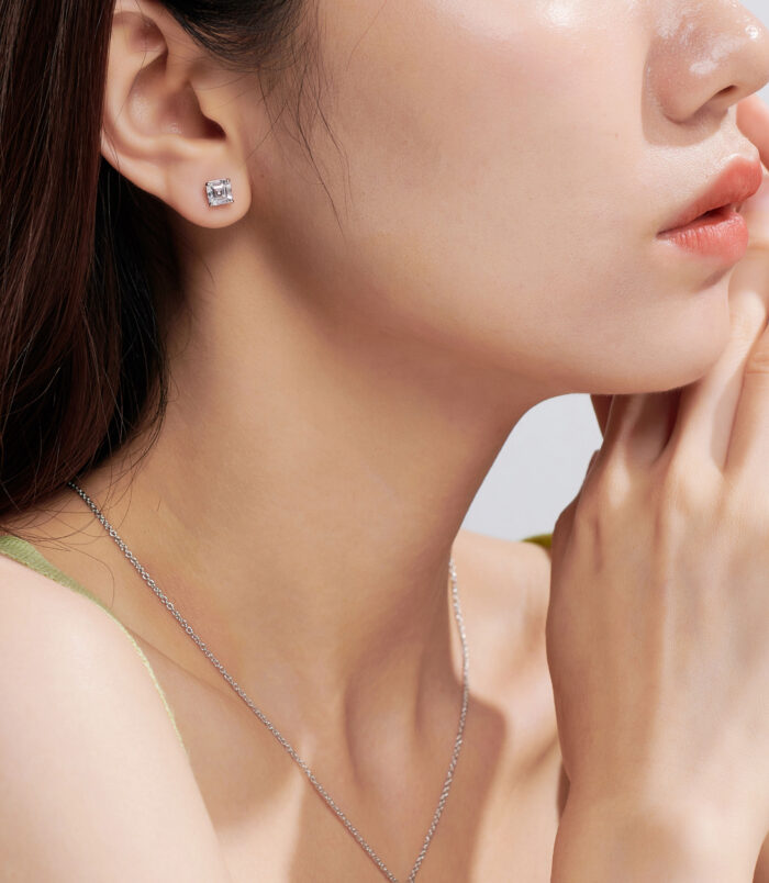 A pair of square, princess-cut stud earrings in polished sterling silver, featuring a central clear gemstone set in a sleek four-prong setting, displayed on a model's ear against a soft, neutral background.