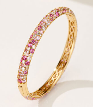 a close-up of a gold bangle bracelet with a gradient of pink and white diamonds, featuring a secure clasp and intricate cut-out pattern on the inner side