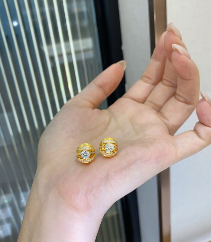 Pair of gold-plated silver stud earrings with a central round stone and halo of small crystals, displayed on a woman’s hand